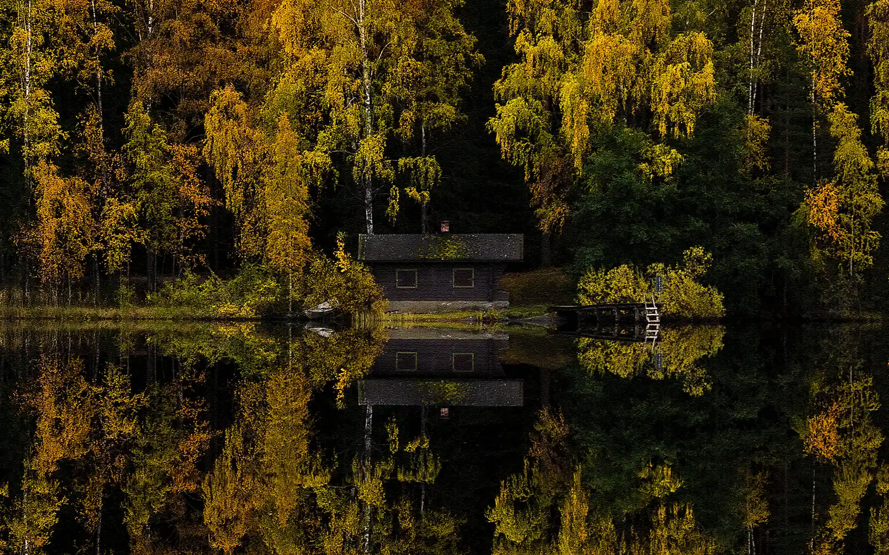 Syksyn kirkkaan keltaista loistoa. Maisema heijastuu peilityynestä veden pinnasta. Keskellä Kintulammin saunarakennus ja laituri.